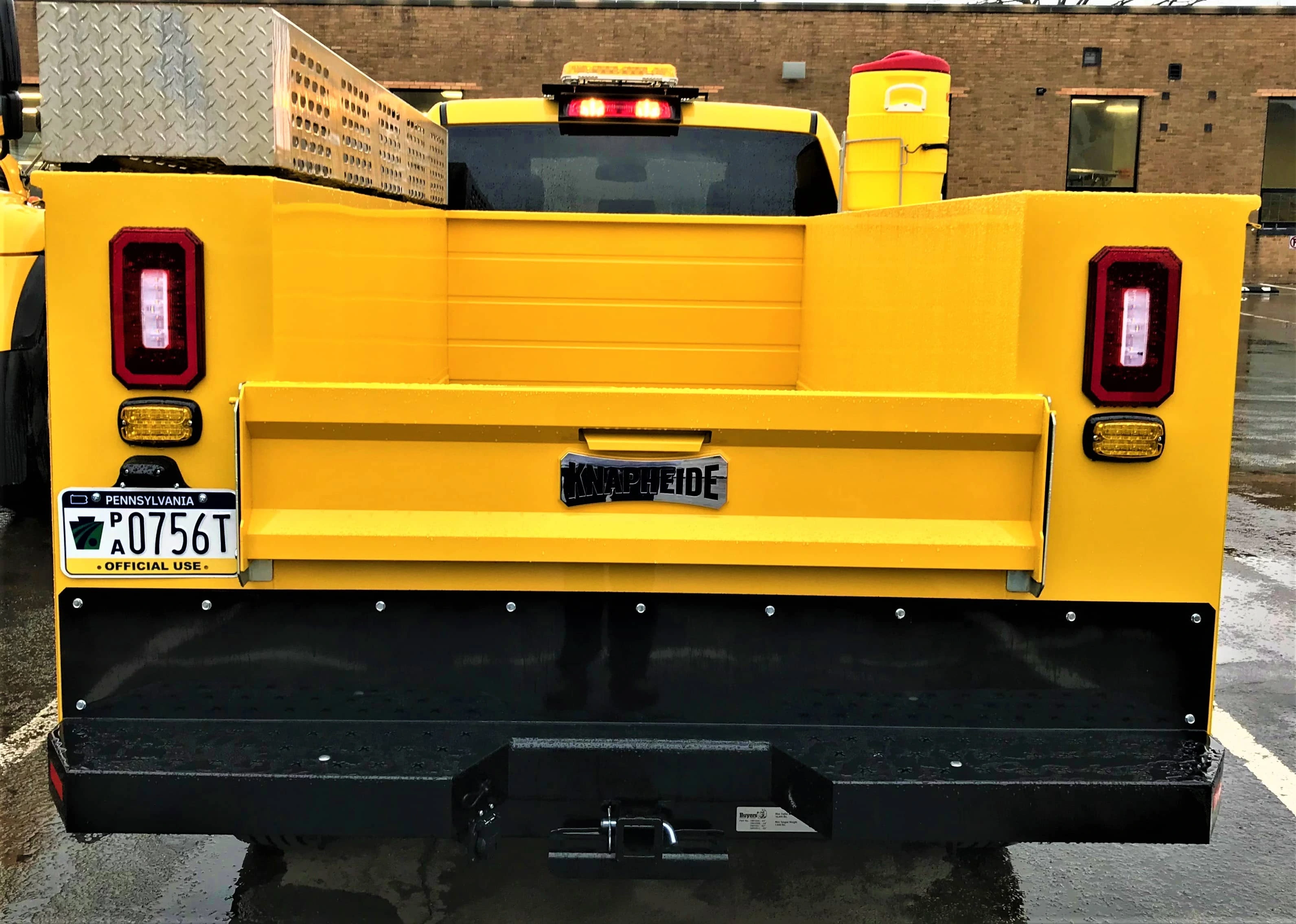 The tailgate of a yellow PennDOT crew cab pick-up truck shows a steel equipment storage box mounted to the top of the left side of the truck bed.